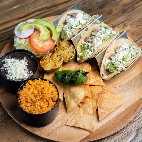 Mexican Mariachi Grill's tacos, rice, beans, salad, chips, grilled lime, and charred pepper on a wooden tray.