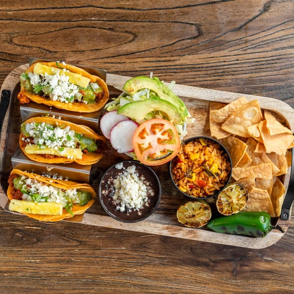 Three Mexican tacos with pineapple, cheese, and salsa, served with rice, beans, chips, and grilled lime.