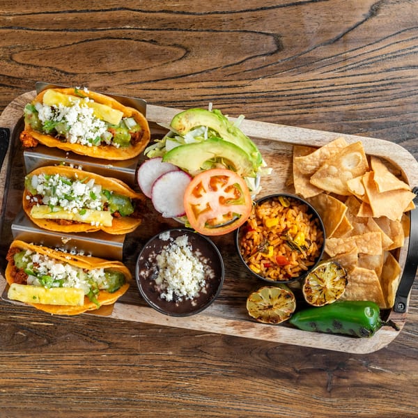 Three Mexican Mariachi Grill tacos with rice, beans, chips, and grilled vegetables on a wooden tray.